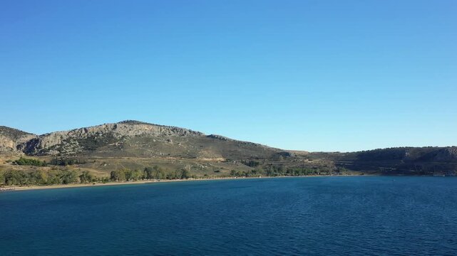Wide view of Paralia Karathona beach with clear blue water and rugged hills under a bright sky near Nafplio, Greece.
