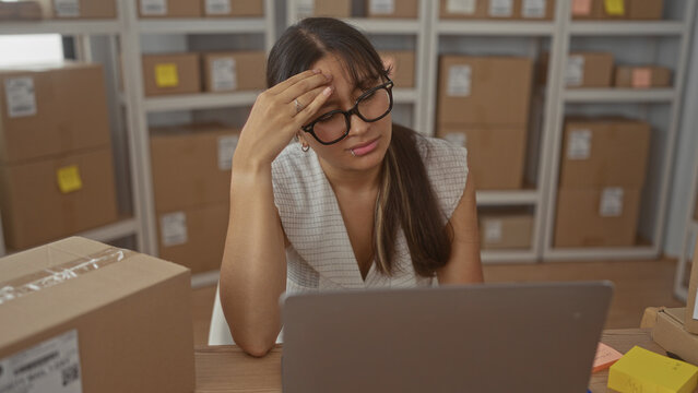 Woman with hand on forehead working on laptop beside parcels and shelves in a storage building; stress small business.