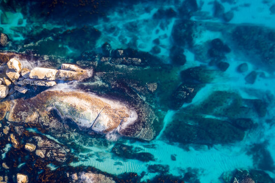 Aerial view of the ocean's turquoise waters gently lapping against the rugged shoreline rocks, creating a mesmerizing dance of light and shadow, Cape Town, Western Cape, South Africa.