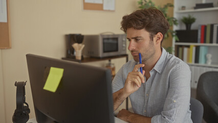 Man holding blue pen to his lips while gazing at a computer monitor in an office workspace with bookshelves and a plant visible  contemplation. © Krakenimages.com