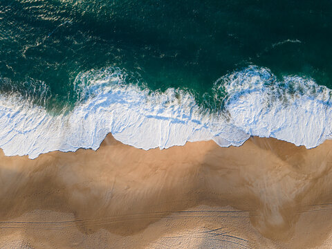 Aerial view of the vibrant turquoise waves crashing onto the golden sands, creating a mesmerizing contrast of color and texture, Nazare, Leiria District, Portugal.