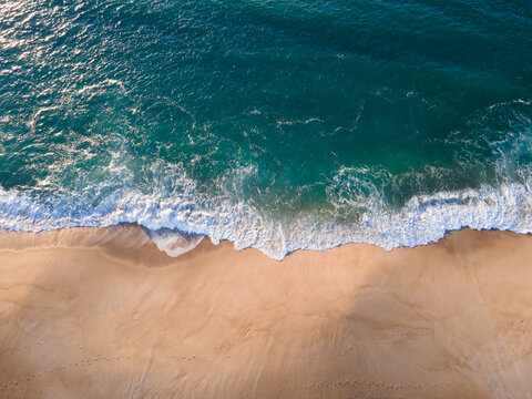 Aerial view of the shoreline where the turquoise waves meet the golden sand, creating a stunning contrast, Nazare, Leiria District, Portugal.