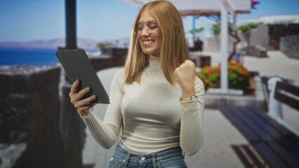 Fototapeta premium Redhead woman pointing finger at tablet and holding device while smiling with a small fist pump on a seaside street promenade; joy success.