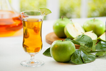 Fototapeta premium Fresh green apple juice in a glass with whole and sliced Granny Smith apples on a wooden table, bright garden background.