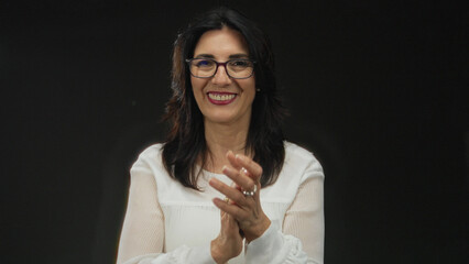 Fototapeta premium Middle-aged hispanic woman claps hands wearing white blouse in black studio; celebration joy encouragement.