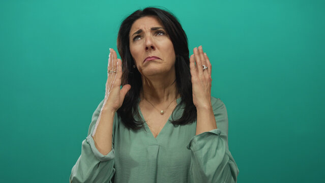 Middle aged woman raises hands to face in teal studio setting with expressive gesture and closed eyes showing despair; frustration.