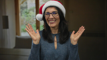 Fototapeta premium Woman with glasses wearing red santa hat waves open palms and smiles against neutral studio backdrop; holiday cheer.