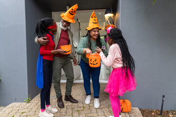 Naklejka premium African American family standing at home doorway holding jack-o'-lantern buckets in pumpkin hats