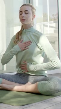 Vertical video: Woman in greentop placing hands on chest and belly breathing calmly on mat by glass