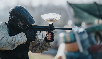 A masked shooter aims a paintball marker in camouflage gear. An outdoor field scene shows players, barriers, and spectators in the background. © qunica.com