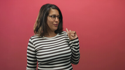 Fototapeta premium Woman points index finger to the right, wearing striped shirt and glasses, brunette middle age portrait in a studio; playful confidence.