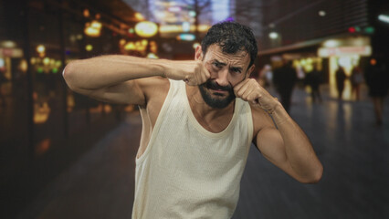 Fototapeta premium Man rubbing eyes with fists in sleeveless shirt on a busy night street with blurred neon shops and pedestrians; urban fatigue.