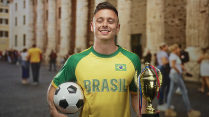 Fototapeta premium Young man in brasil jersey holding soccer ball and trophy on a street with stone columns; victory pride.