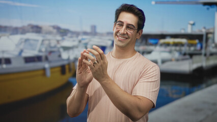Fototapeta premium Man claps hands and smiles in a studio set staged as a marina by a dock with boats visible, wearing glasses and rings on his fingers, midgesture capturing motion; contentment.