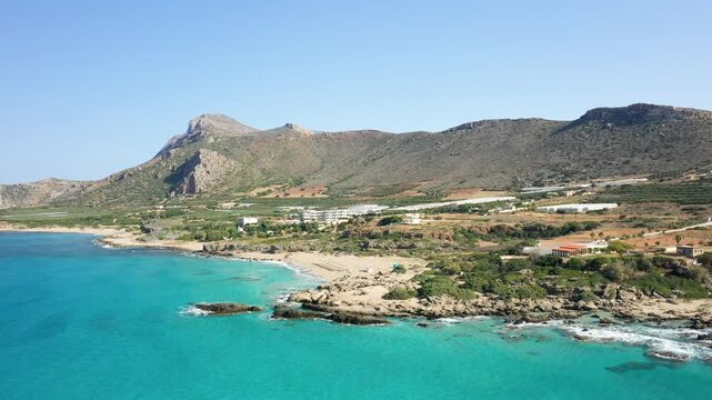 Scenic aerial view of Falasarna Beach with turquoise waters, rocky shoreline, and mountains in Crete, Greece under a clear blue sky.