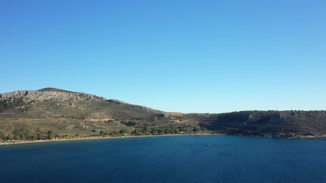 Wide view of Karathona Beach with clear blue water and rocky hills under a cloudless sky in Nauplie, Greece. Natural coastal landscape ideal for travel and tourism concepts.