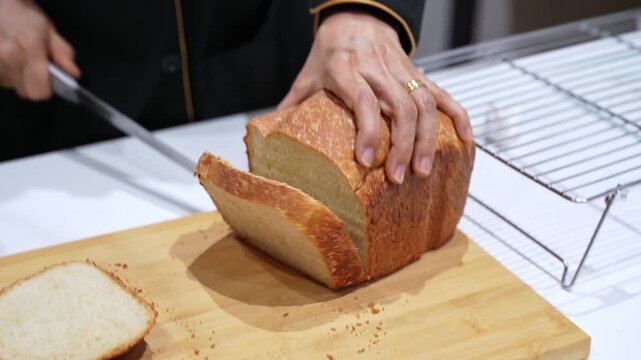 Close-up of hands cutting a fresh homemade bread loaf with a serrated knife on a wooden board, crumbs on table beside a cooling rack in a bright kitchen.