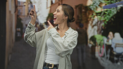 Fototapeta premium Woman pointing finger upward and smiling in a narrow street cafe alley, striped shirt visible; joy curiosity.