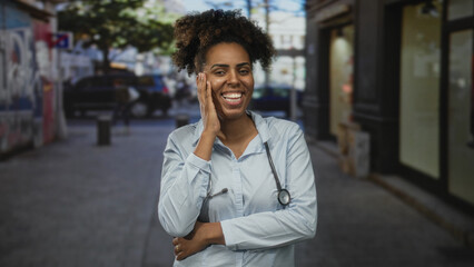 Woman doctor with stethoscope smiling, hand to cheek on street with urban shops and parked cars; community care confidence.