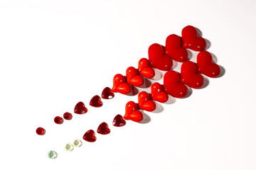 Glossy red and orange heart-shaped ornaments arranged diagonally on a white background, with a trail of small faceted heart gemstones leading toward them. Minimal romantic concept with copy space.