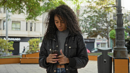 Woman with smartphone scrolling and typing with both hands in street near bench and lamppost,...