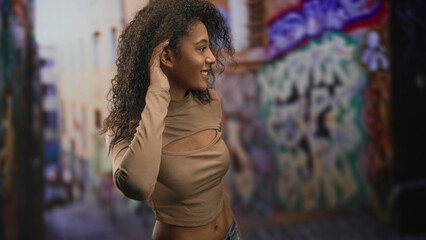 Obraz premium Teenage woman with curly hair holds hand to ear and covers mouth in surprise, midriff visible against graffiti wall on a street; joyful.