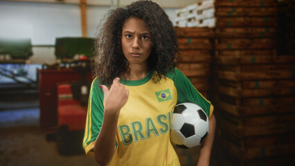 Teenage woman with curly hair wearing yellow brazil jersey holds soccer ball under arm and points...