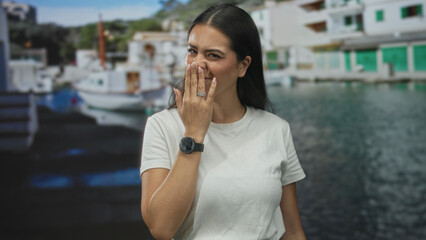 Fototapeta premium Woman in white t shirt covers mouth with hand while smiling and squinting on street by boats and water; amusement.