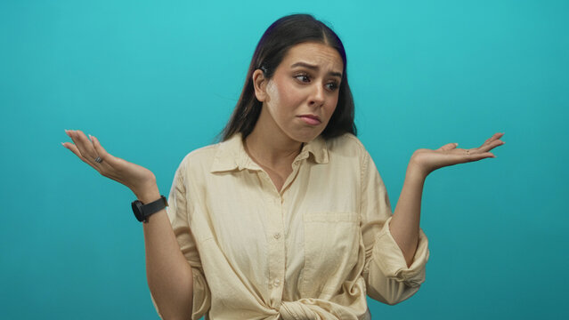 Young brunette woman shrugging with palms up and hands visible in teal studio setting, wearing beige shirt and smartwatch; uncertainty dilemma.