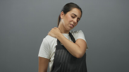 Obraz premium Woman in striped apron with hand on shoulder, eyes closed and grimacing suggesting neck strain in gray studio; discomfort.