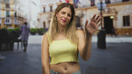 Young woman in yellow crop top showing bare midriff, standing and waving in a sunlit city street...