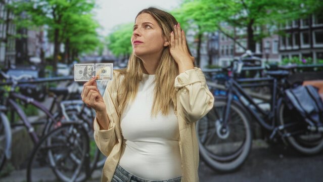 Woman holding a us dollar banknote in her right hand and touching chin while standing on a street with bicycles; financial uncertainty doubt.