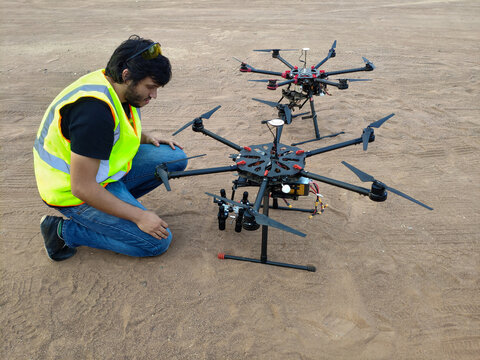 technician man stands with two professional hexacopter UAVs sitting on sand inspecting the parts