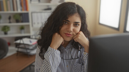 Woman adjusting glasses with hand, slight smile while seated at desk by monitor and bookshelves in an office building  confidence. © Krakenimages.com