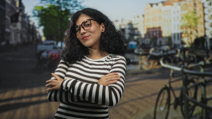 Woman with arms crossed on a street in amsterdam by canal and bicycles, wearing striped top and...