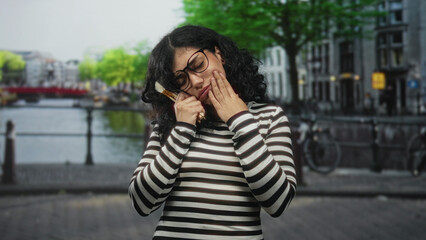 Fototapeta premium Arab woman with glasses and striped sweater holding makeup brushes, hand to chin on street in amsterdam; creative doubt.