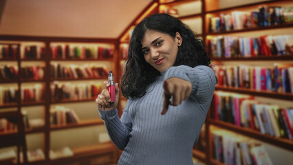 Young woman holding vape and pointing finger forward in a library building filled with bookshelves and warm light  youth leisure confidence. © Krakenimages.com