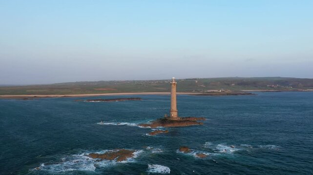 A tall lighthouse stands on a rocky islet surrounded by the sea, with the coastline of Cap de la Hague, Normandy visible in the background under a clear sky.