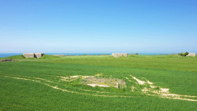 Wide view of lush green fields with historic concrete bunkers under a clear blue sky near the coast at Longues sur Mer, Normandy, France.