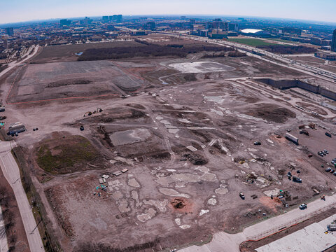 Aerial view of a vast expanse of cleared land, marked by patches of exposed earth and scattered debris, contrasts sharply with the distant skyline of modern buildings, Frisco, Texas, United States.