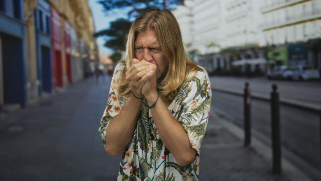 Blond man with long hair covers nose with hand on city street sidewalk near bollards amid empty storefronts; disgust.