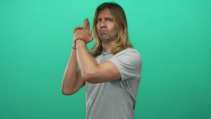 Fototapeta premium Man with long blond hair pointing finger like gun with stern puckered lips in green studio; playful mischief.