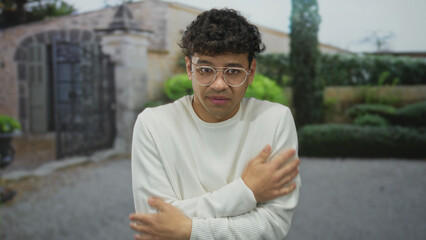 Fototapeta premium Young man hugs bare shoulders outside stone building entrance as he shivers in light sweater; anxiety.