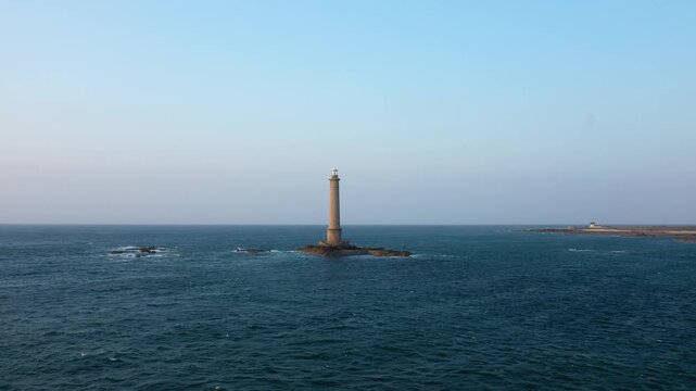 Solitary lighthouse stands on a rocky islet surrounded by the blue sea under a clear sky at Cap de la Hague, Normandy, France. Tranquil maritime landscape with coastal horizon.