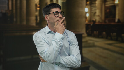 Obraz premium Young hispanic man in a light blue shirt touches lips with finger inside church building; quiet contemplation.
