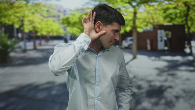 Hispanic man in white shirt cups ear on a sunlit city street lined with green trees and parked cars; curiosity.