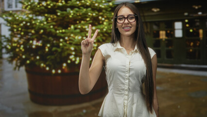 Fototapeta premium Teenage girl with glasses and braces points index finger then flashes peace sign beside a decorated christmas tree on a street; festive cheerful.