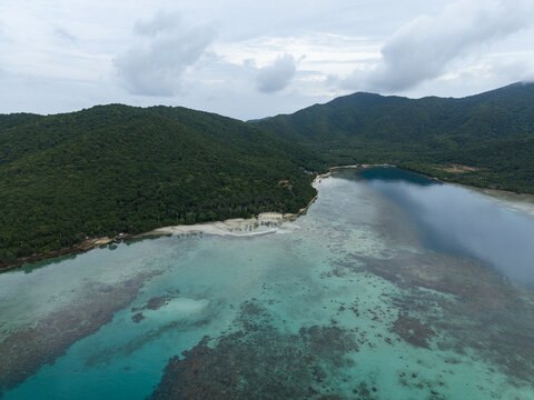 Aerial view of turquoise waters meeting verdant, hilly landscapes under a cloudy sky at Bobby Beach, Karimunjawa, Central Java, Indonesia.