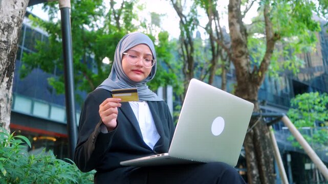 Portrait of a stressed Asian Muslim businesswoman wearing hijab, using a laptop and credit card, having payment issues outdoors against a modern office building and green background.