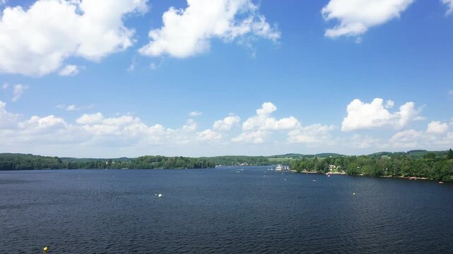 Wide view of Lac des Settons with calm water, green shoreline, and a bright blue sky dotted with fluffy white clouds. A small boat creates gentle ripples on the peaceful lake.